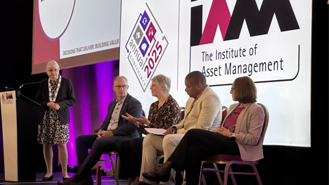 Five people on stage at an IAM event; one stands at a podium with the IAM logo, and a screen behind displays 